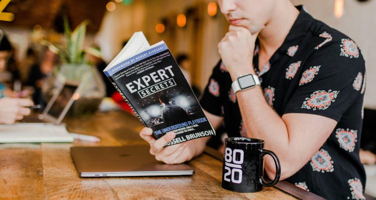 An image of a young man reading a book titled Expert Secrets.